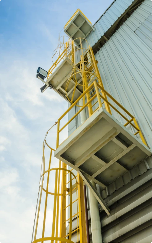 Escada marinheiro amarela com gaiola de proteção e patamares, instalada na lateral de estrutura industrial metálica.