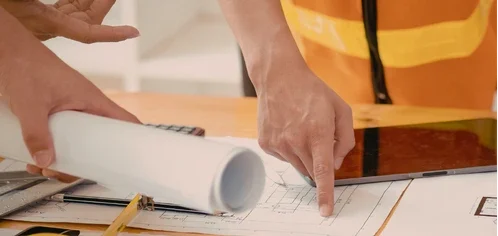 Mãos analisando planta técnica com rolo de projeto sobre mesa, representando planejamento e engenharia.