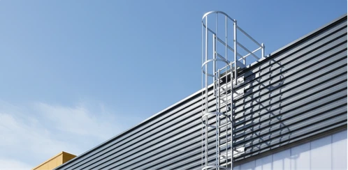 Escada marinheiro fixada na fachada metálica de um galpão, com guarda-corpo no topo sob céu azul.