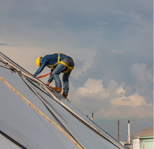 Profissional com cinturão de segurança realizando manutenção em telhado inclinado, preso a sistema de ancoragem.