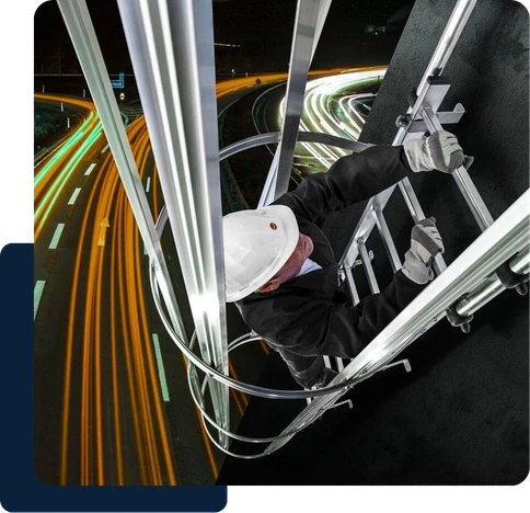 Técnico com capacete instalando escada marinheiro com linha de vida em estrutura metálica, com vias iluminadas ao fundo.