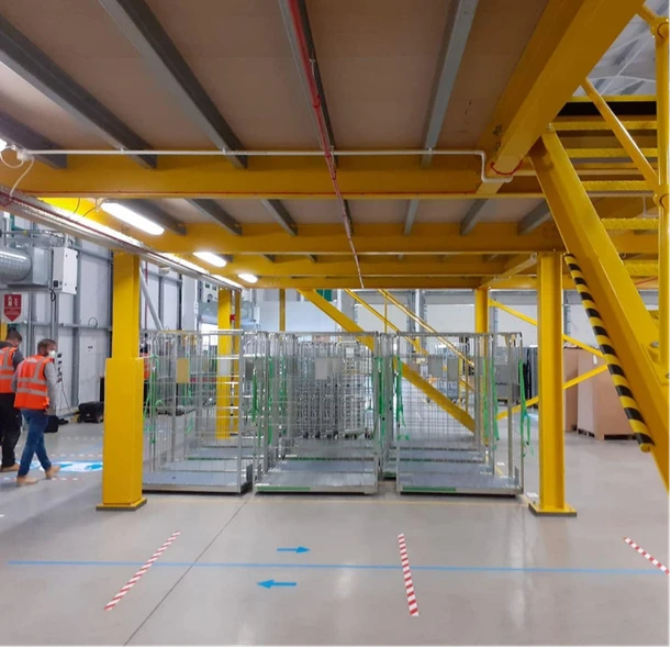 Mezanino estrutural industrial com pilares amarelos e escada de acesso em galpão logístico com sinalização de segurança.